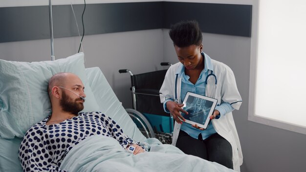 Специалист-хирург чернокожая женщина-врач объясняет диагноз болезни, показывая рентгеновский снимок с помощью планшетного компьютера в больничной палате. Больной пациент, отдыхающий в постели, обсуждает лечение выздоровления