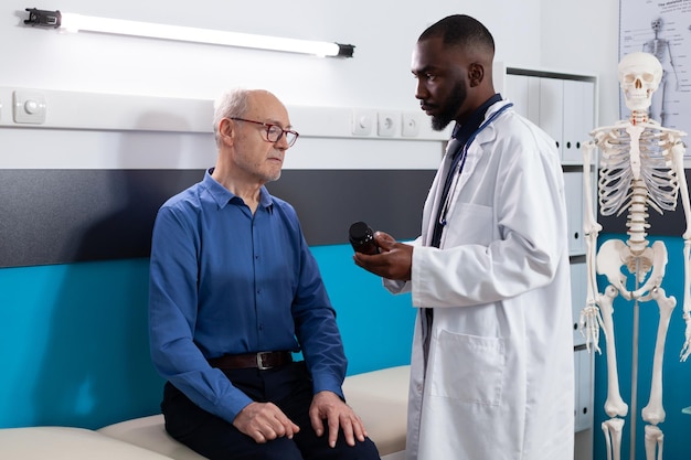 Specialist therapist doctor holding pills bottle explaining medication treatment to retired old man discussing disease symptoms during medical consultation in hospital office. Medicine concept