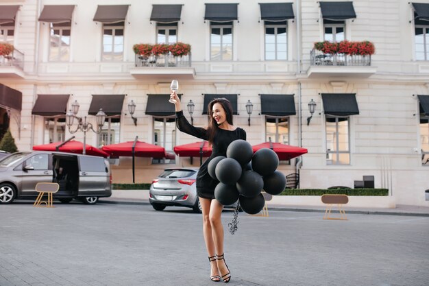 Free Photo spectacular black-haired lady raising glass of wine, while posing on the street early in evening