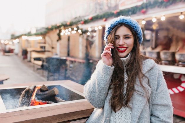 笑顔でクリスマスフェアでポーズウールのグレーのコートで壮大なブルネットの女性。長い髪型のロマンチックな女の子は、冬の休日のために装飾された通りに立っている青い帽子をかぶっています。