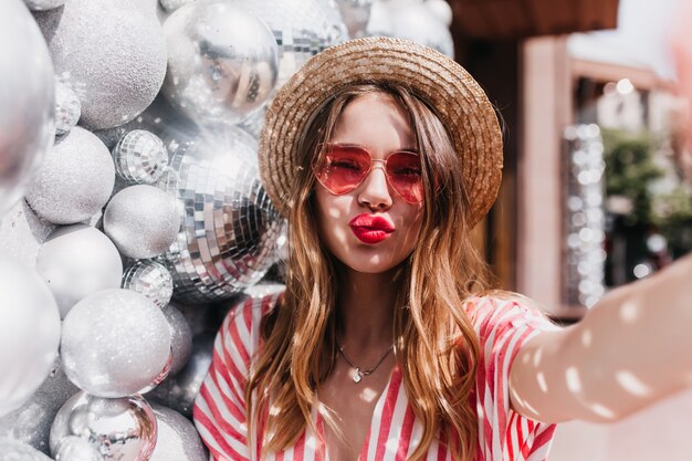 Spectacular female model in good mood making selfie with kissing face expression. Outdoor photo of debonair blonde woman wears summer hat standing near sparkle balls.