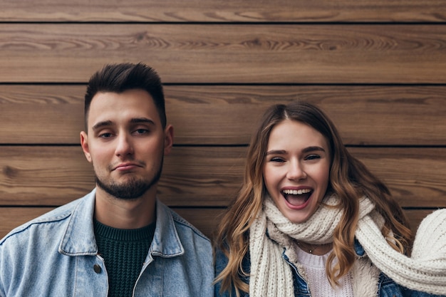 Spettacolare ragazza in abito autunnale che si gode il servizio fotografico con il fidanzato. foto interna di due amici in posa con il sorriso sulla parete in legno.