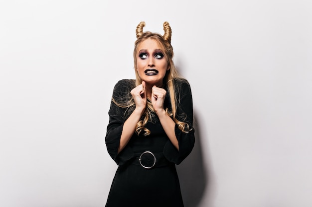 Spectacular girl in dress posing during halloween photoshoot. Funny scared lady in witch costume standing on white wall.