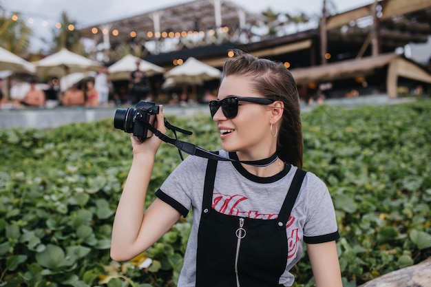 Foto gratuita spettacolare ragazza con capelli castani tenendo la fotocamera vicino a prato verde. foto all'aperto della donna europea attraente che fa le foto nel parco.