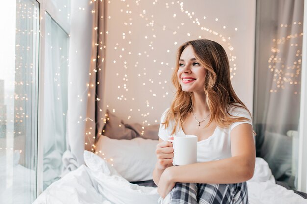 Spectacular girl with wavy hair looking at window while drinking tea. Indoor shot of lazy blonde woman enjoying morning in bed.