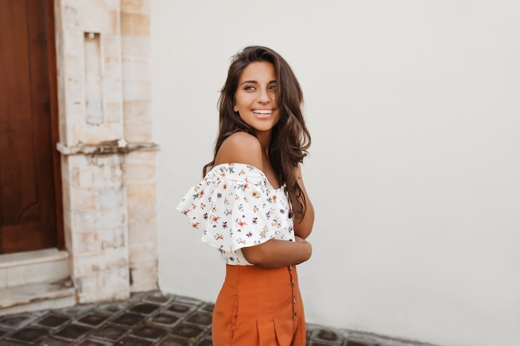 Free Photo spectacular lady with wavy dark hair and tanned skin poses against white building with wooden door