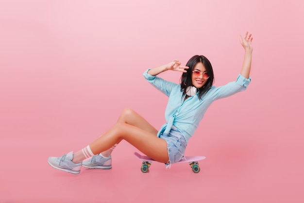 Spectacular latin girl in cute outfit sitting on skateboard and waving hands with happy face expression. laughing hispanic young woman in sunglasses and trendy sneakers.