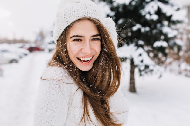 Эффектная длинноволосая женщина смеется, позируя на снегу. Наружное фото крупным планом кавказской женской модели с романтичной улыбкой охлаждает в парке в зимний день.
