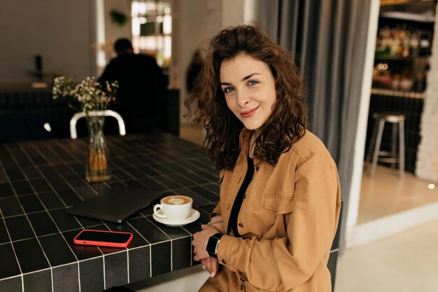 Spectacular lovable girl with curls dressed brown shirt is looking at camera with wonderful smile while sitting in cafe with coffee