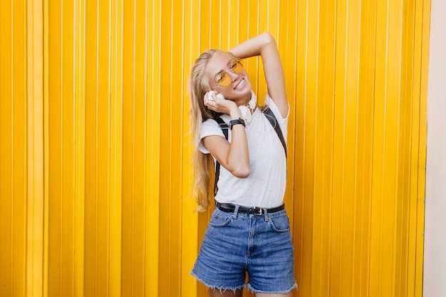 Spectacular slim woman holding white headphones. Outdoor portrait of blonde carefree girl posing on yellow background.