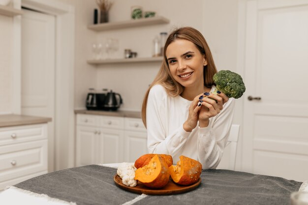 Эффектная улыбающаяся дама сидит на кухне с брокколи и тыквой, готовится к приготовлению