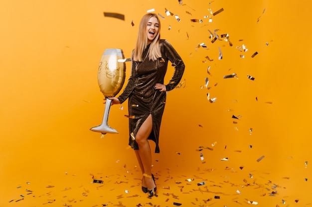 Spectacular tall woman posing with toy wineglass. Laughing blinde woman in party dress dancing on orange.