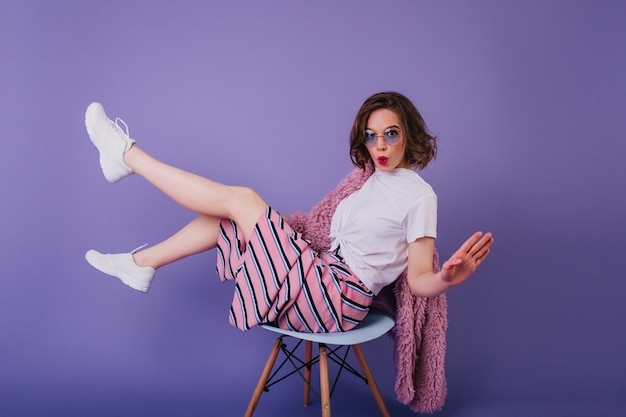 紫色の壁に浮かんでいるサングラスの壮大な白人の女の子。椅子に座っている流行のスニーカーでかなりヨーロッパの女性モデル。