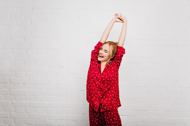 Spectacular woman in trendy sleepwear stretching with smile. Enthusiastic girl enjoying good morning and posing on white wall.