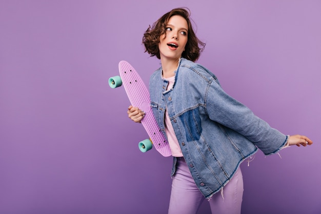 Spectacular young lady in oversize jacket dancing. Portrait of dreamy curly girl with skateboard.