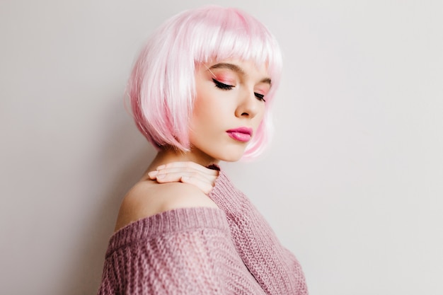 Spectacular young lady in stylish periwig standing with eyes closed. Pretty white girl wears pink wig posing in soft purple sweater.