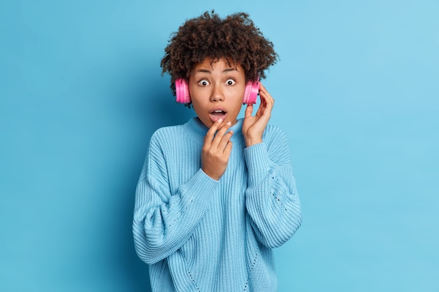 Speechless dark skinned young Afro American woman has impressed face expression wears stereo headphones keeps mouth opened dressed in oversized knitted sweater 
