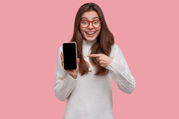 La giovane donna eccitata senza parole indica la schermata di imitazione dello smartphone, cerca di mostrare qualcosa di fantastico, indossa un maglione bianco