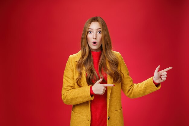 Speechless questioned and confused surprised redhead girl in yellow autumn coat pointing right open ...