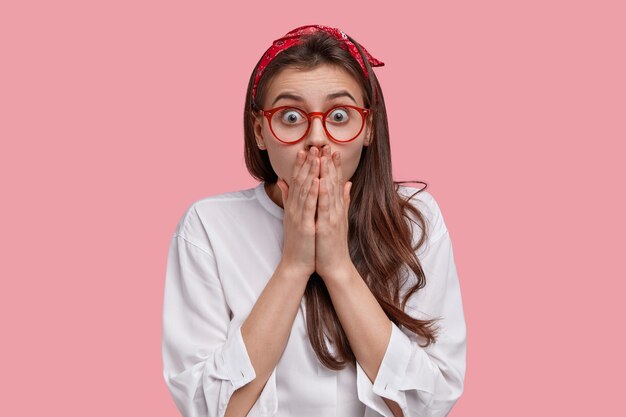 Speechless young woman stares with astonishment, covers mouth with both hands, looks perplexed, wears spectacles, headband and white shirt