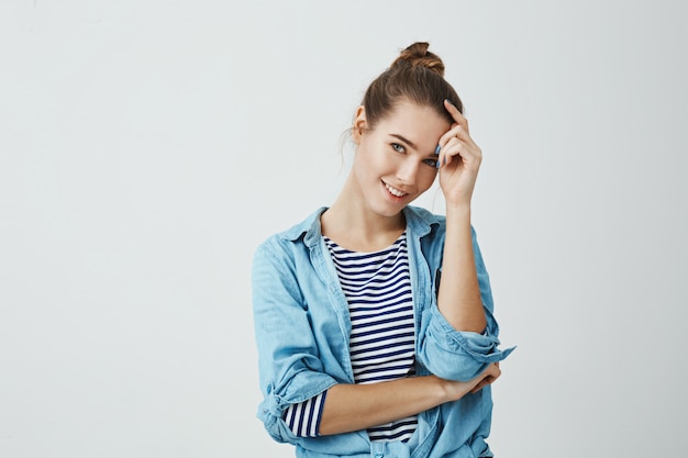 Spend life on bright side. Indoor shot of attractive young caucasian student with bun hairstyle, holding hand on forehead while tilting head and smiling , being shy .