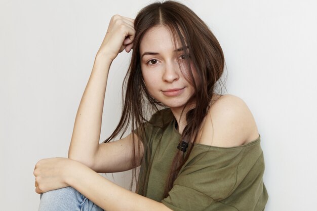 Spending home alone. Portrait of stylish teenage woman wearing dark messy hair in braid looking with cute smile, leaning elbow on her knee. Young European woman with pretty face posing indoors