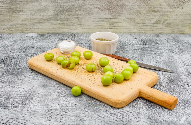 Пряные зеленые сливы в разделочной доске с небольшим кусочком соли, сушеным тимьяном и фруктовым ножом, вид сбоку на поверхность гранж и деревянную стену