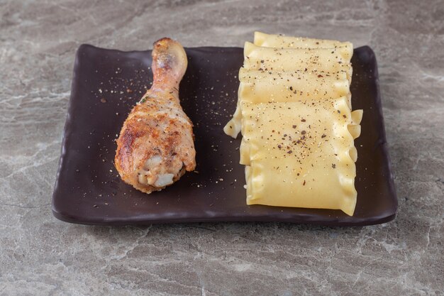 Spicy lasagna sheets alongside chicken drumstick on the wooden plate, on the marble.