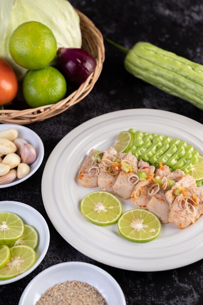 Spicy lime pork salad with bitter melon, galangal, chili, tomatoes and garlic in a white plate on a black cement floor.