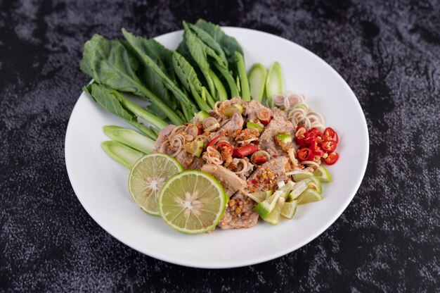 Spicy lime pork salad with kale, galangal, chilli and garlic in a white plate on a black cement floor.