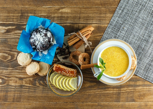 Spicy milk in a cup with mint, biscuits, cloves, lemon slices, cinnamon sticks top view on wooden table