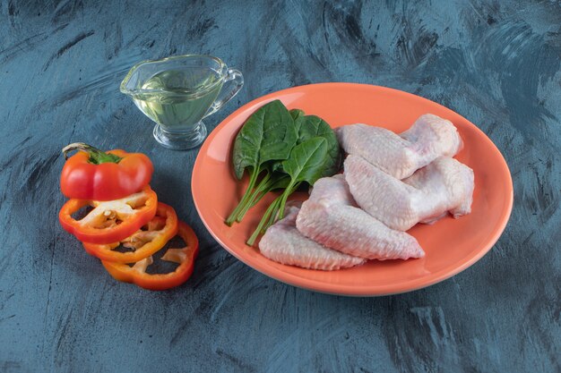 Шпинат и куриное крылышко на тарелке рядом с нарезанным перцем и миской масла на синей поверхности.
