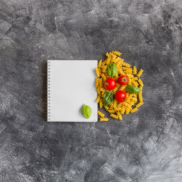 Spiral notebook with raw fusilli; tomatoes and basil leaves