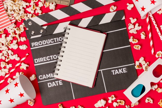 Spiral notepad on clapperboard with disposable glass; popcorns; drinking straw and 3d glasses on red backdrop