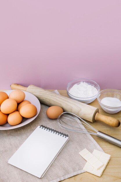 Spiral notepad; eggs; whisk; rolling pin; sugar; white chocolate and flour on desk