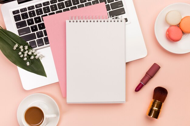 Спиральный блокнот на ноутбуке с миндальным печеньем и кофейной чашкой с кисточками для макияжа на цветном фоне