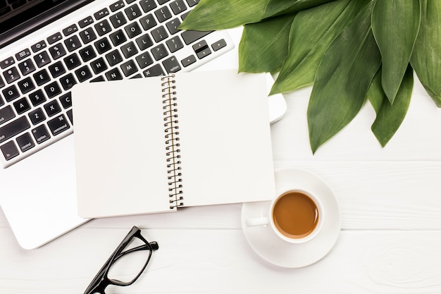 Foto gratuita blocco note a spirale e foglie sul computer portatile con gli occhiali e tazza di caffè sulla scrivania in legno