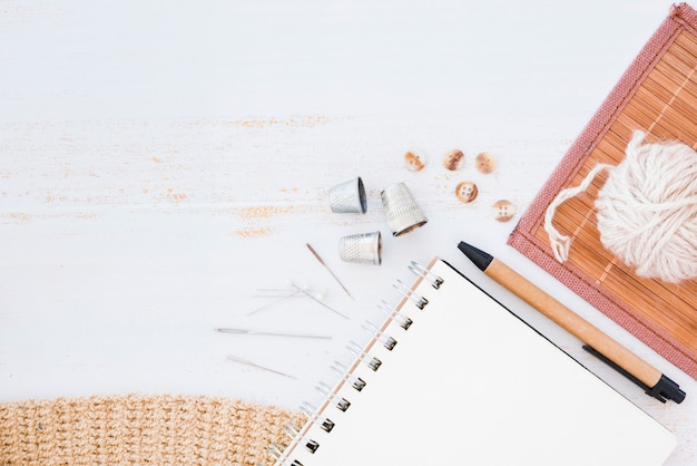 Spiral notepad; needles; thimble; buttons; pen; wool ball on placemat over wooden textured backdrop