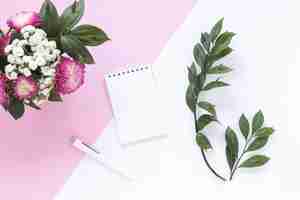 Free photo spiral notepad; pen; leaves and bunch of flowers on dual background