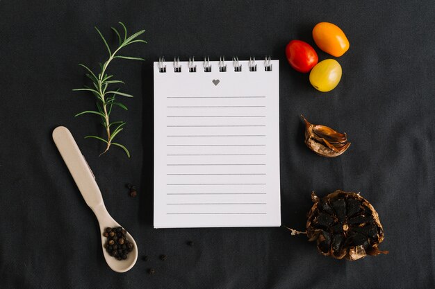 Spiral notepad surrounded with rosemary; cherry tomatoes; black pepper and garlic