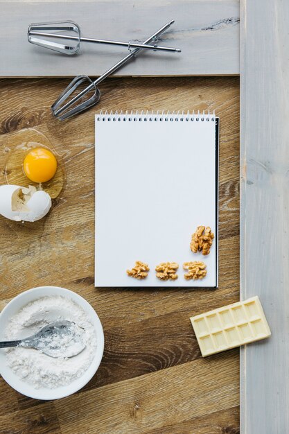 Spiral notepad; walnut; chocolate; flour; egg and whisk on wooden surface