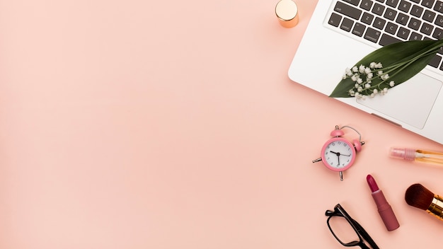 Free photo spiral notepad with lipstick,alarm clock,eyeglasses on laptop against peach backdrop