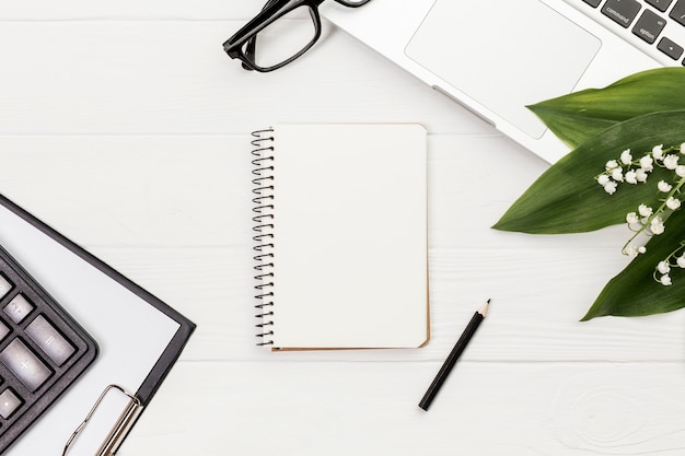 Spiral notepad with pencil,calculator,clipboard,eyeglasses and laptop on white desk