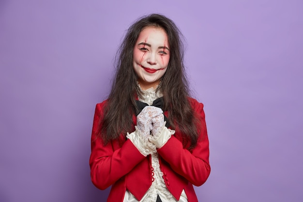 Spooky happy woman has image of zombie keeps hands together wears ghost makeup poses for halloween poster isolated on purple wall