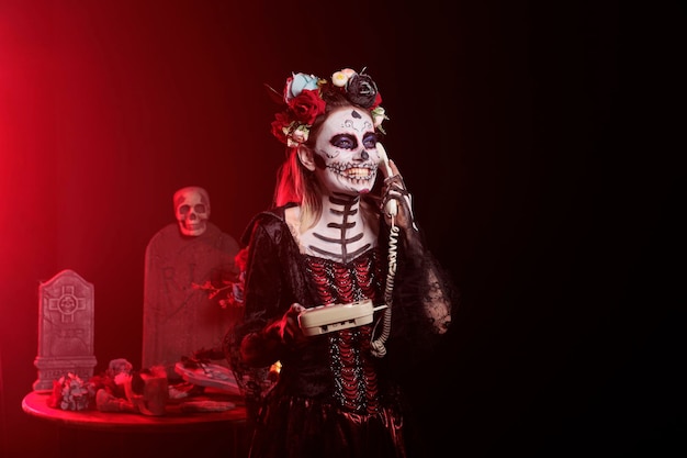 Foto gratuita donna spettrale che risponde al telefono fisso con cavo, parla al telefono dell'ufficio mentre ha il trucco del cranio e la body art. raffigurante la signora della morte di santa muerte nel giorno della celebrazione del giorno dei morti.