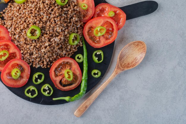 Spoon next to a wooden board with buckwheat, pepper and tomato on marble surface