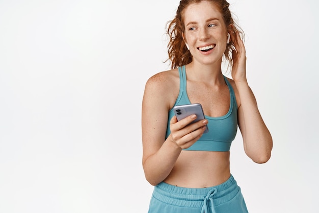 Sport and fitness Smiling female athlete listening music in headphones holding mobile phone standing in activewear against white background