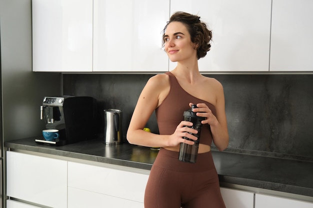 Free photo sport and healthy lifestyle portrait of smiling fitness woman in activewear standing near kitchen co