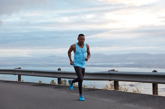 Sport, motivation and recreation concept. Active male sporty black jogger runs against cloudless sky in highway, wears casual vest and blue sportshoes, has biceps on arms, exercises outdoor.