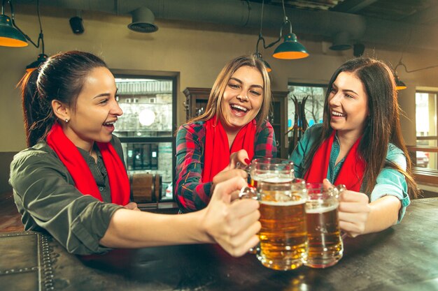 Free photo sport, people, leisure, friendship, entertainment concept - happy female football fans or good young friends drinking beer, celebrating victory at bar or pub. human positive emotions concept